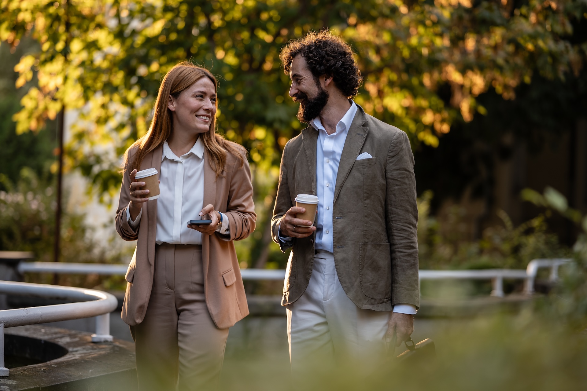 Business partners walking outdoors, holding disposable coffee cups and discussing work during a coffee break
